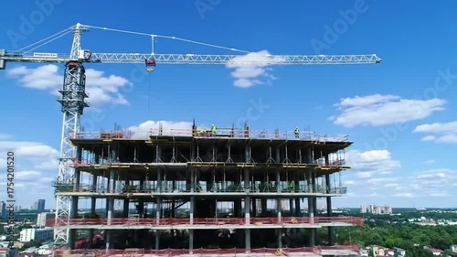 Construction site with a towering building and large white crane against a bright blue sky with scattered clouds