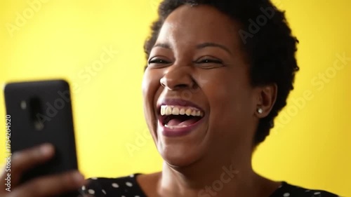 A cheerful Black woman laughing exuberantly at her phone, with a bright yellow background