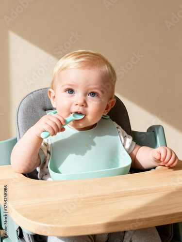 Blonde toddler sitting in high chair eating independently with silicone bib and spoon in bright natural home setting