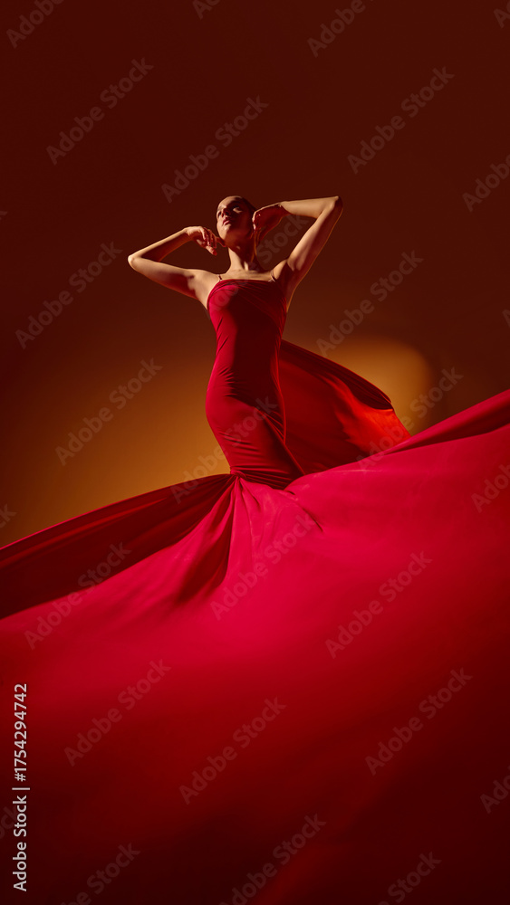 Fototapeta premium Female model in red dress posing under warm studio light with flowing fabric. Concept of fashion style, elegance, color harmony for editorial, commercial, and design projects.