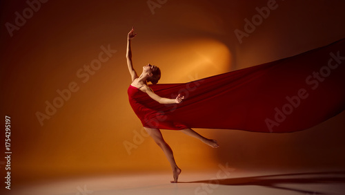 Ballet dancer in red fabric leaping under warm orange spotlight in studio. Concept of movement, artistic performance, and professional dance photography for commercial use.