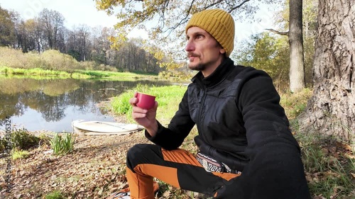 A man in a bright yellow beanie enjoys a ripe apple by a peaceful riverside in autumn. The vibrant scenery with colorful leaves and tranquil waters captures natures beauty, bringing serenity
