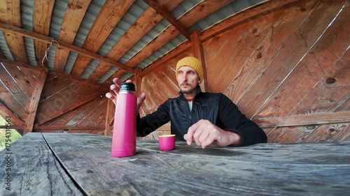 A man in a yellow beanie and black jacket is enjoying pouring a hot drink from a pink thermos into a small cup at a rustic wooden table in a cozy cabin, relishing the warmth around him