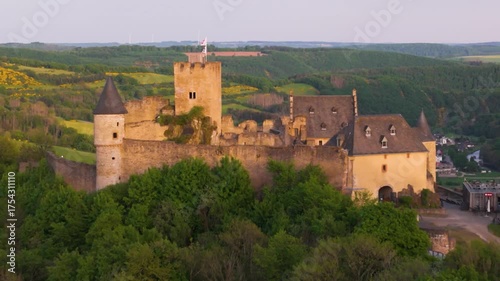 bourscheid castle château de green valley cover hills behind walls towers windmills hilltops cloudy sky town below from toward afar forward drone shot 