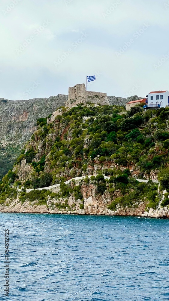 Naklejka premium Vertical photo of Kastellorizo Megisti, Greece. Ferry view of colorful houses by the sea with mountains in background. Mediterranean island, coastal landscape, summer vacation, travel, scenic seaside