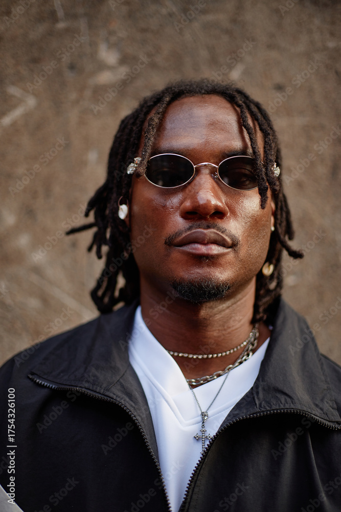 Fototapeta premium Portrait of young adult Black man wearing sunglasses and jewelry looking directly at camera standing against textured wall background, displaying confident facial expression
