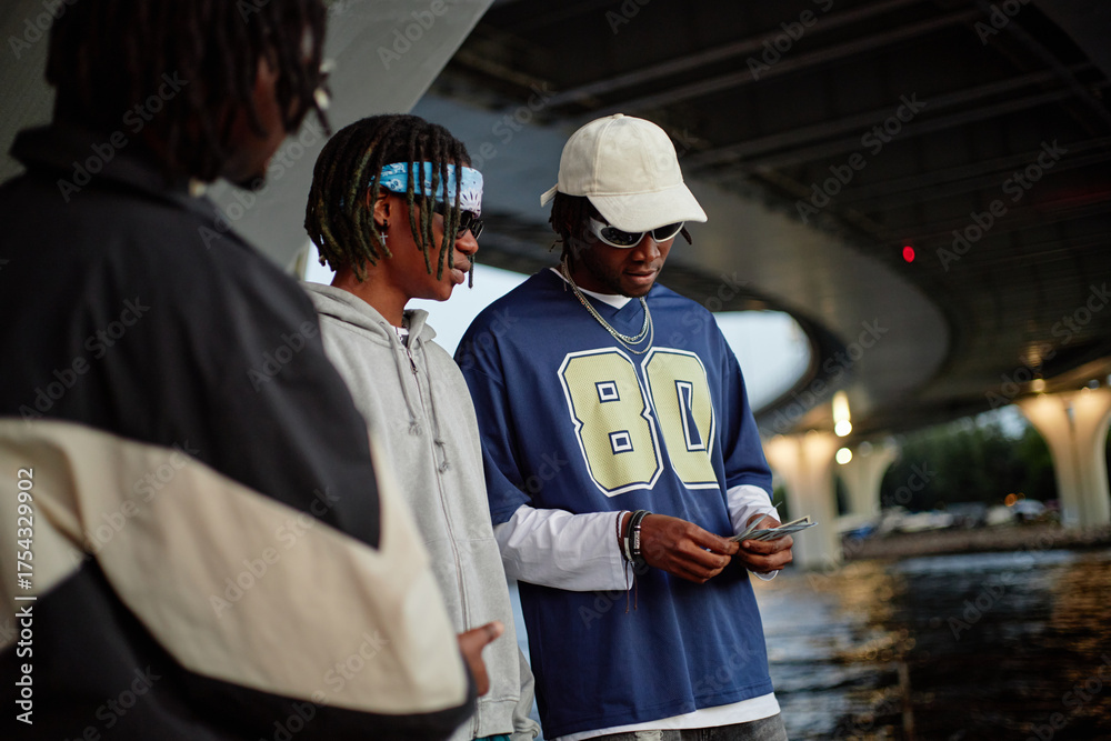 Obraz premium Three Black men standing under bridge near water, one wearing cap and sunglasses counting money while others watching, casual outdoor urban setting