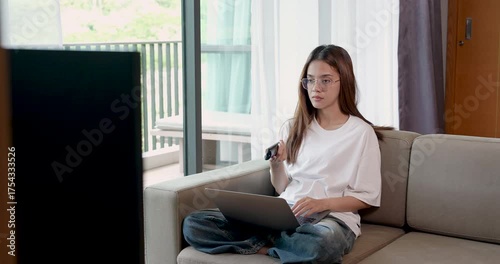 Asian adult woman sits cross-legged on sofa working remotely on laptop, feeling mentally tired and bored from repetitive home office tasks under soft daylight in quiet modern living space