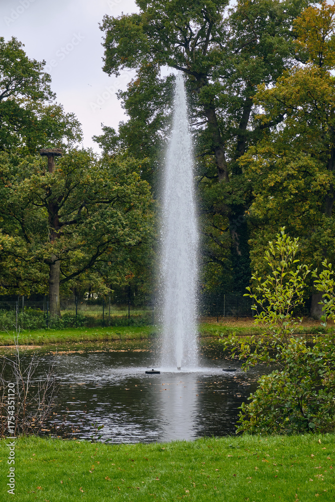 Naklejka premium Fountain in the large pond of the Nienoord estate in Leek, Netherlands, surrounded by autumn foliage on a rainy October day, reflecting the calm and classic atmosphere of this historic landscape
