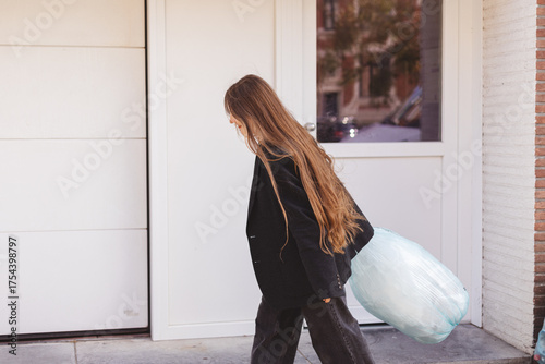 Woman taking out the trash, holding a garbage bag outdoors near recycling bins before work. Concept of daily routine, waste management, eco lifestyle, responsibility, cleanliness and sustainability.