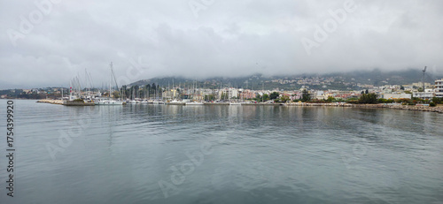 A serene harbor scene with sailboats docked along the shore, surrounded by misty mountains and cloudy skies