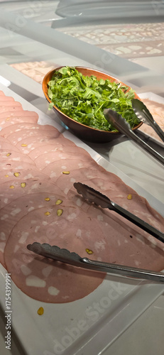 Sliced mortadella with pistachios on a platter with salad and tongs.