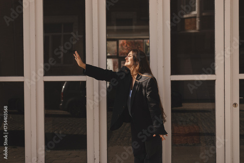 Young professional woman in a stylish blazer smiles and waves outdoors in warm sunlight. Modern urban lifestyle portrait conveying confidence, friendliness, fashion forward business energy, optimism.