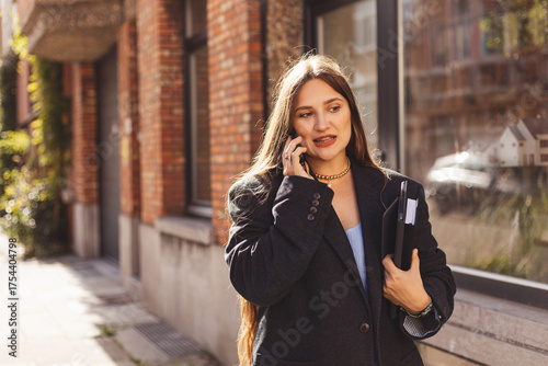 Stylish businesswoman walking in the city, talking on smartphone and holding folder with documents. Modern urban lifestyle concept showing communication, confidence, and professional attitude.