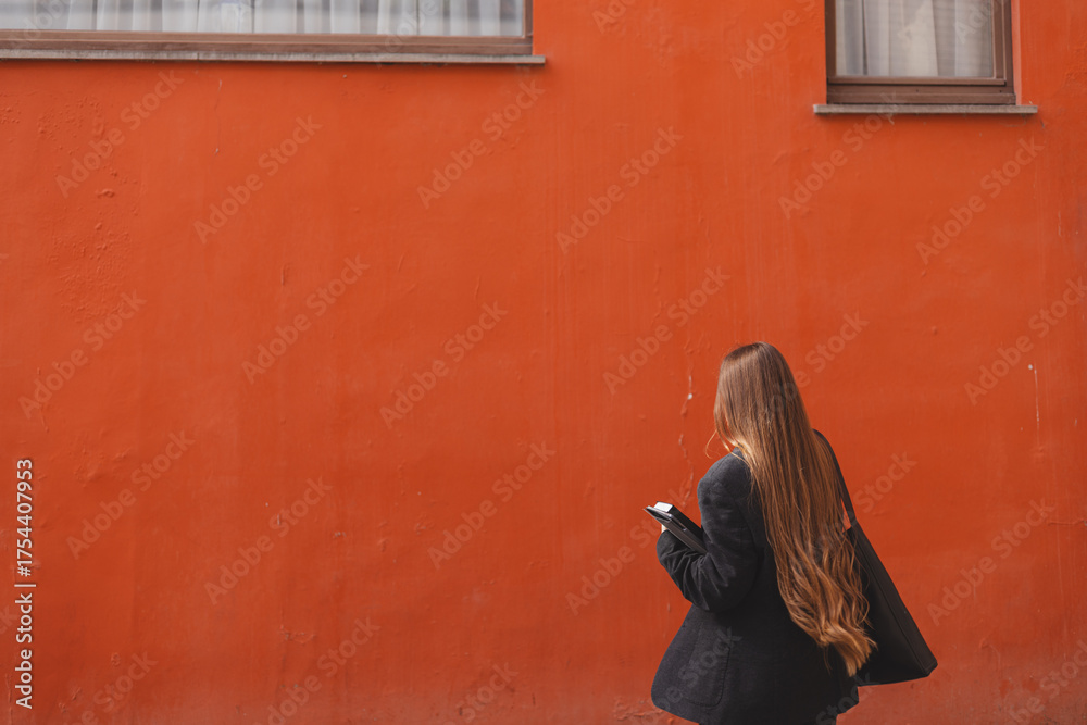 Naklejka premium Serious young adult business woman walks and talks on phone, solves problems, holds documents, notebook, tablet and bag on background of orange wall. Girl dressed in black jacket, denim jeans. Back.
