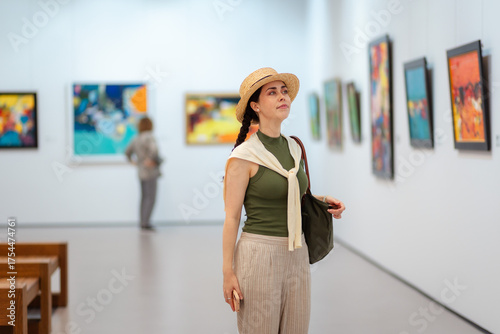 Mid shot of young pretty Caucasian woman in a straw hat and elegant clothes visits a contemporary art gallery. Paintings in the background. Concept of Museum Day