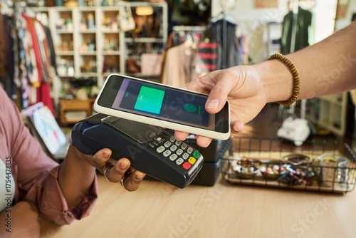 Caucasian young adult man holding smartphone over payment terminal making contactless payment, while Black woman holding card reader in thrift shop with clothing and accessories in background