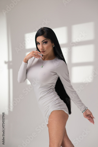 Stylish brunette woman in gray short dress posing indoors. Soft window light casts geometric shadows, creating a minimalist, editorial mood. Studio comercial corporate photography