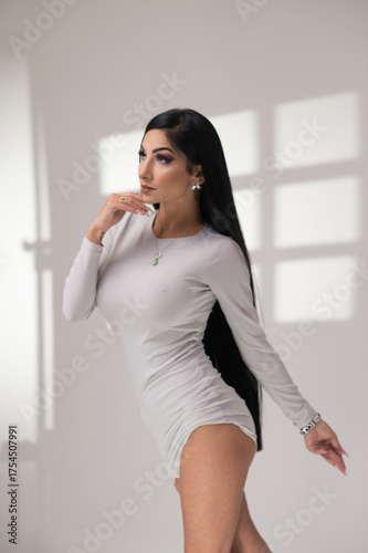 Stylish brunette woman in gray short dress posing indoors. Soft window light casts geometric shadows, creating a minimalist, editorial mood. Studio comercial corporate photography