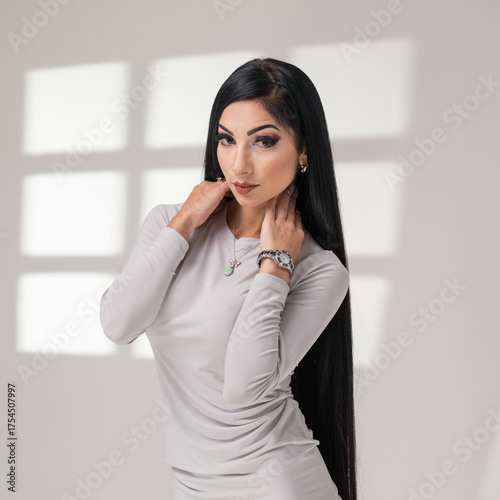 Stylish brunette woman in gray short dress posing indoors. Soft window light casts geometric shadows, creating a minimalist, editorial mood. Studio comercial corporate photography