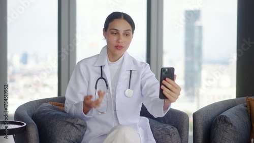 Young woman doctor is enjoying a friendly video call on her mobile phone with a patient or friend while sitting comfortably on a sofa. Female cheerful demeanor adds warmth to the cozy living room