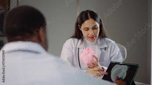 Two cardiologists engage in a detailed discussion on heart disease treatment, using an anatomical model to illustrate their points while consulting with a medical doctor in a clinic, share knowledge