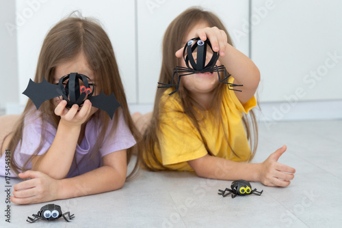 Two little sisters lying on the floor and playing with Halloween themed paper crafts they made themselves. Creative kids activity and holiday preparation at home.