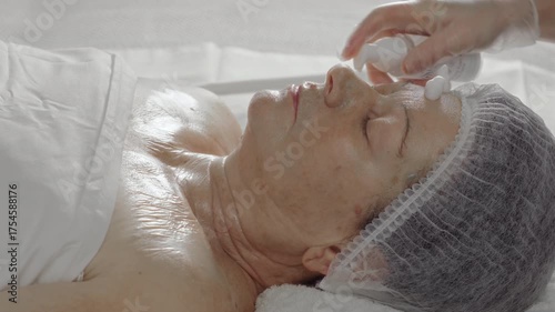 A cosmetologist applies an alginate mask wash-off product to the face of an elderly woman