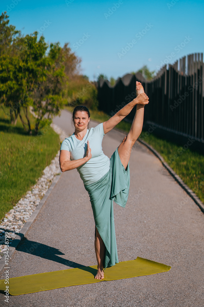 Naklejka premium Woman striking an elegant standing yoga pose on a sunlit path, exemplifying flexibility, focus, and grace in harmonious outdoor setting.