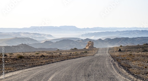 Gravel track leads through barren desert landscape, sandstone rock formation, silhouettes of mountains against the light, Damaraland, Kunene, Namibia