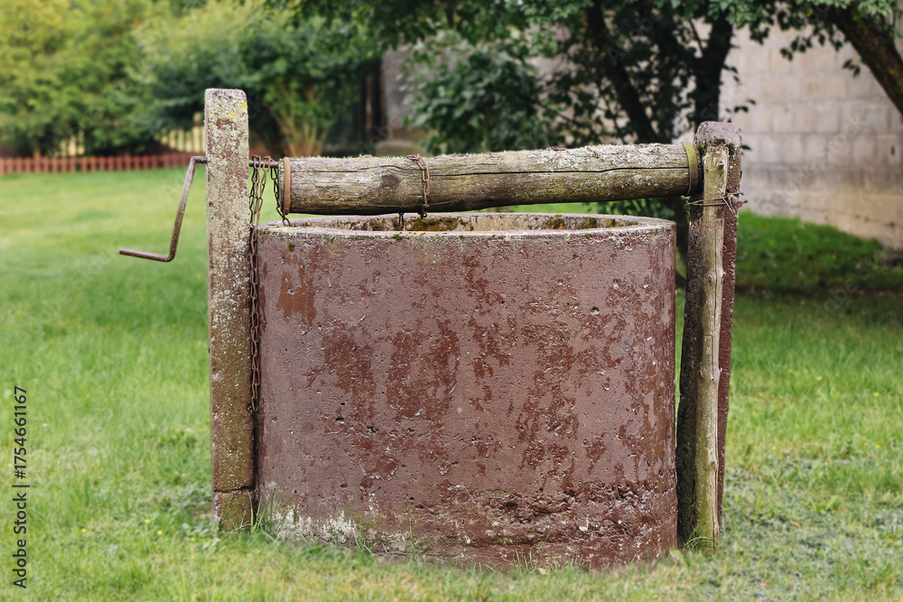 Fototapeta premium Old rustic water well in green garden. Traditional countryside construction with metal cylinder and wooden handle showing history and simplicity of rural life.