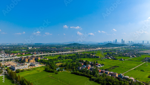 Aerial view of lush green fields and rural houses with a modern city skyline in the distance.