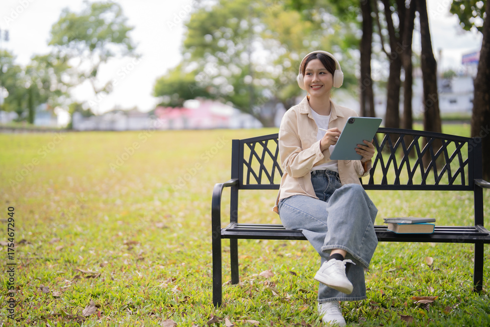 Fototapeta premium Young Asian woman wearing headphones and using a digital tablet with a stylus while sitting on a park bench, engaging in e-learning in a serene outdoor environment