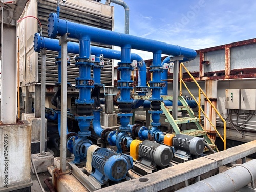 An intricate industrial cooling system on a rooftop featuring electric pumps, a network of blue pipes, flanges, and valves. This HVAC and plumbing machinery highlights heavy industry and utility maint