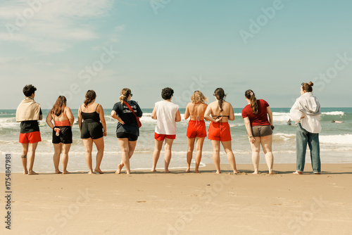 Grupo de amigos de pie en fila en la playa, observando las olas del océano en un día soleado de verano.