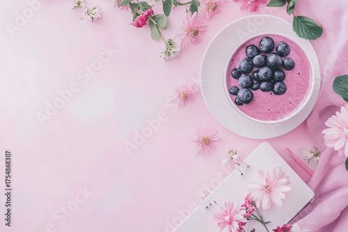 Pink smoothie bowl with fresh blueberry topping surrounded by pink flowers and notebook on soft pink background creating calm and delicate atmosphere