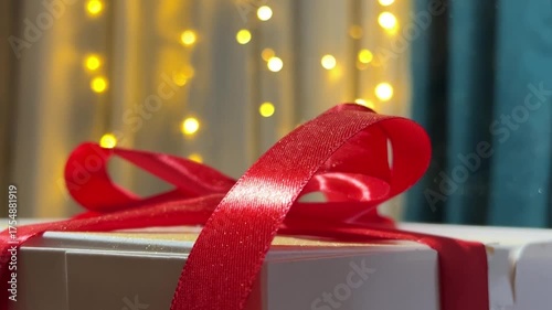 Close up video. 60 fps. Magic dust falling on white gift box with red bow, satin ribbon on garland background. Gold yellow lights bokeh. Concept of Happy New Year, Merry Christmas, birthday holiday