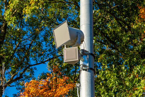photo of a exterior stadium speakers typically found at a sporting event, mounted on a galvanized light pole, with trees in the background.  
