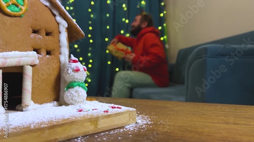 4K defocused video. 60 fps. Lovers, friends, spouses gives New Year gifts. Man with beard out of focus sitting on blue sofa in room. Gingerbread brown house with snowman in foreground. Winter decor