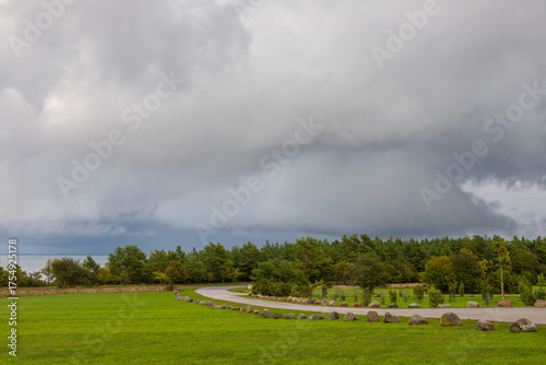Curved road through green field under dramatic cloudy sky, Estonia.