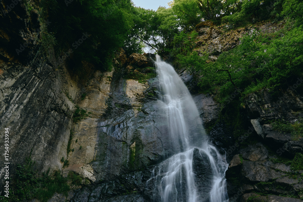 Obraz premium Majestic waterfall cascading down rocky cliffs surrounded by lush green forest, creating a serene and natural landscape full of movement and freshness.