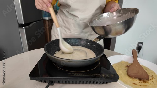 A woman makes pancakes, pours batter into a frying pan with a ladle from a bowl