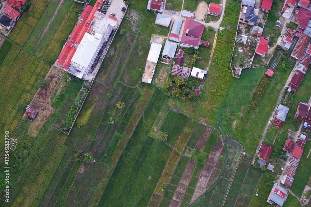 Fototapeta premium Aerial view of flooded rice fields, irrigation canals, and expanding urban settlements in Sungai Penuh, Jambi, Indonesia. Showing urban sprawl, farmland conversion, and environmental landscape.