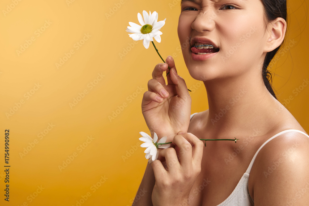 Fototapeta premium asian woman holds a delicate flower near her face, smiling softly against a warm yellow backdrop, conveying a fresh and gentle mood that highlights natural beauty and calm confidence.