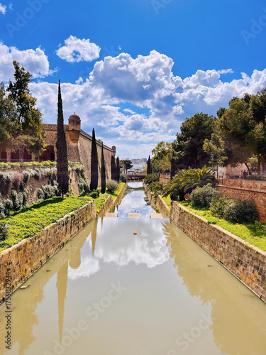 Historische Festungsmauer mit Wassergraben und Zypressen unter blauem Himmel in Palma de Mallorca, Spanien. Malerische Aussicht mit Wolken und Spiegelung im Wasser.