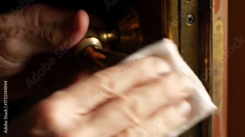 Close-up of a person's hands using a white cloth to clean and disinfect a brass doorknob.