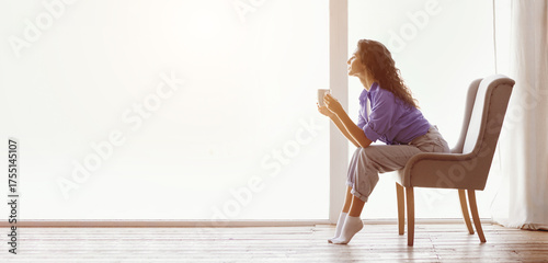 A woman sits quietly in a chair, sipping coffee. She enjoys the peaceful atmosphere and warm sunlight streaming through the window, creating a cozy indoor space.