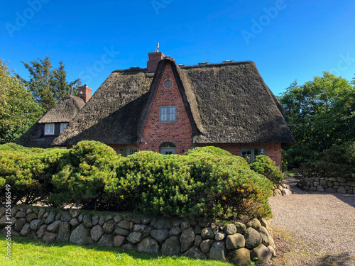 Traditionelles Reetdachhaus aus rotem Backstein, umgeben von Natursteinmauer und grünen Büschen unter blauem Sommerhimmel. Norddeutsche Idylle im typischen Friesenstil.