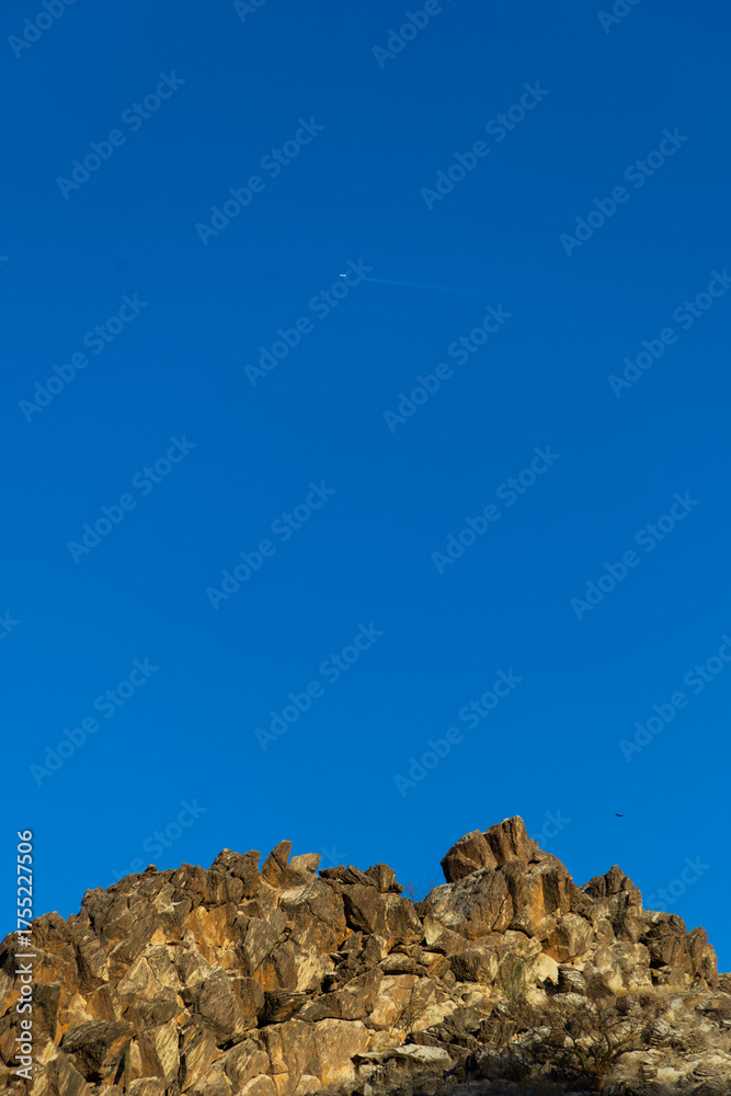 Naklejka premium A summer landscape features a rocky stone wall beneath a blue sky and a passing airplane.
