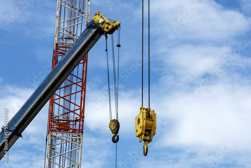 Crane arms extend into midday sky. Red lattice tower frame and blue boom hang empty hooked hoist blocks in open air, waiting for next loads at busy construction site.